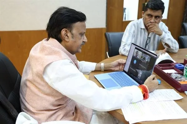 मेज पर लैपटॉप देखते दो अधिकारी, स्क्रीन पर बैठक का दृश्य